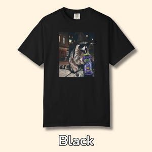 May include: A black t-shirt featuring a graphic print of a raccoon wearing a witch hat and black sunglasses, holding a purple can with a straw. The raccoon is positioned in an outdoor setting at night with blurred city lights in the background.