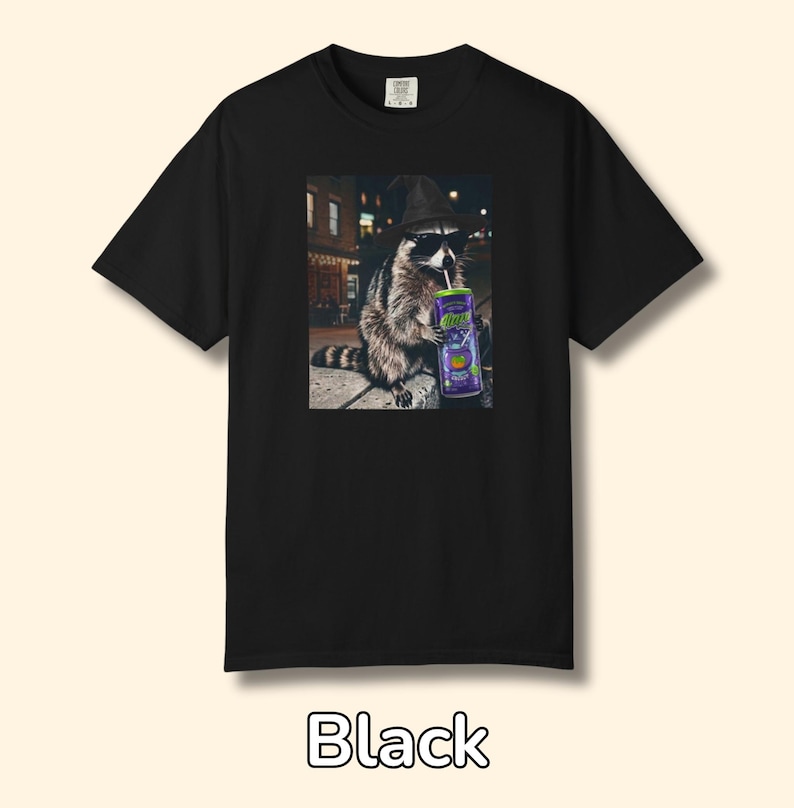 May include: A black t-shirt featuring a graphic print of a raccoon wearing a witch hat and black sunglasses, holding a purple can with a straw. The raccoon is positioned in an outdoor setting at night with blurred city lights in the background.