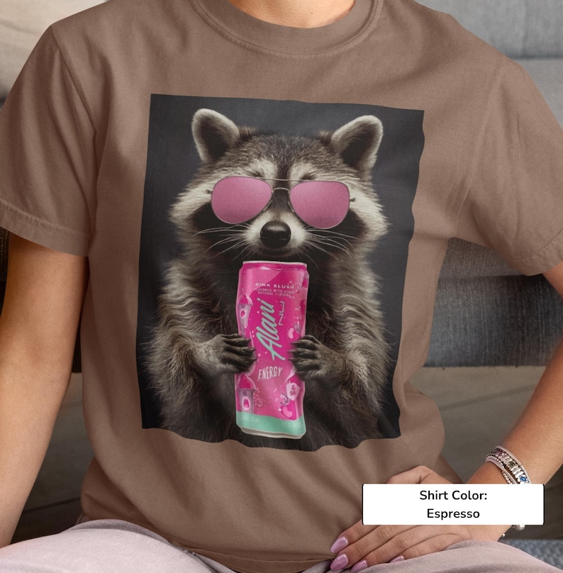 May include: An espresso-colored t-shirt featuring a graphic of a raccoon wearing pink-tinted sunglasses and holding a pink Alani energy drink can. The raccoon is depicted in a realistic style against a dark background, centered on the chest.