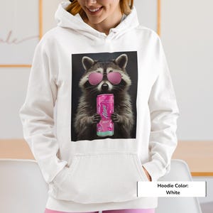 May include: A white hoodie featuring a graphic print of a raccoon wearing pink sunglasses and holding a pink Alani energy drink can. The raccoon is centered on the chest area of the garment, which includes a hood and a front kangaroo pocket.