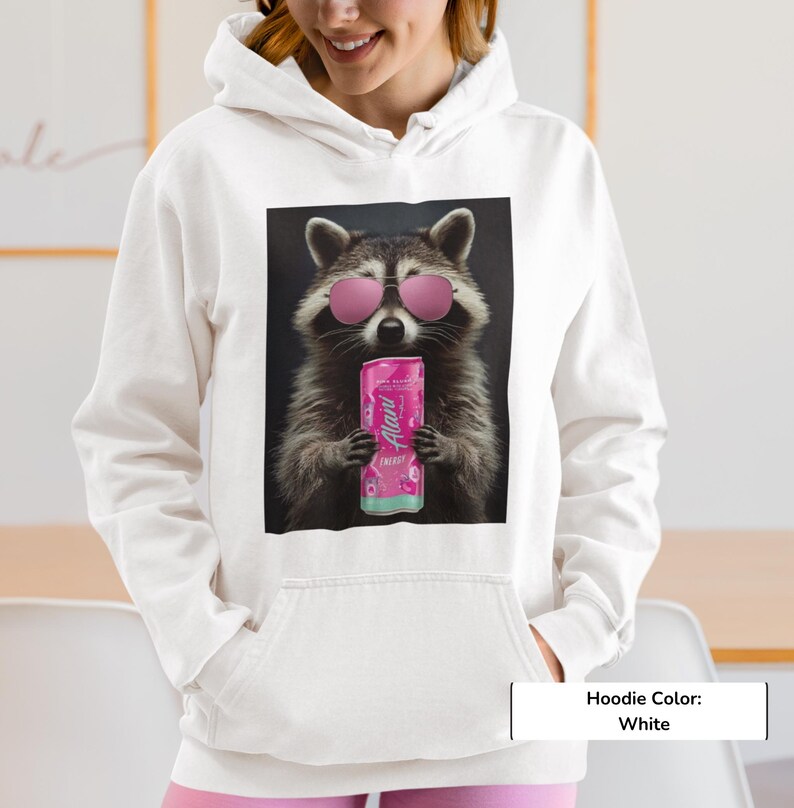May include: A white hoodie featuring a graphic print of a raccoon wearing pink sunglasses and holding a pink Alani energy drink can. The raccoon is centered on the chest area of the garment, which includes a hood and a front kangaroo pocket.