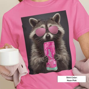 May include: A neon pink t-shirt featuring a graphic print of a raccoon wearing pink-tinted sunglasses and holding a pink Alani Nu energy drink can. The raccoon has grey and black fur and is centered on the front of the shirt against a dark background.