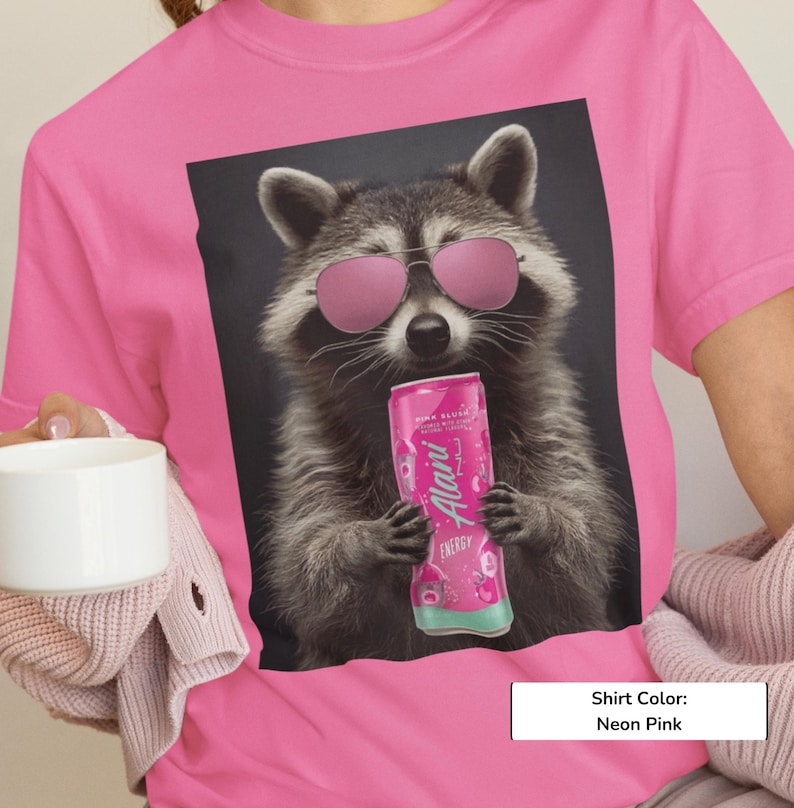 May include: A neon pink t-shirt featuring a graphic print of a raccoon wearing pink-tinted sunglasses and holding a pink Alani Nu energy drink can. The raccoon has grey and black fur and is centered on the front of the shirt against a dark background.
