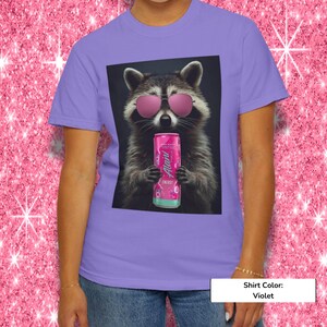 May include: A violet t-shirt featuring a graphic print of a raccoon wearing pink sunglasses and holding a pink Alani Nu energy drink can. The raccoon is centered on the shirt, which has a crew neckline and short sleeves, showcasing a vibrant design.