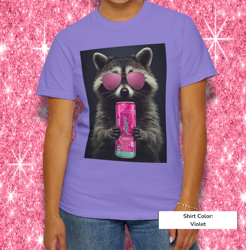 May include: A violet t-shirt featuring a graphic print of a raccoon wearing pink sunglasses and holding a pink Alani Nu energy drink can. The raccoon is centered on the shirt, which has a crew neckline and short sleeves, showcasing a vibrant design.