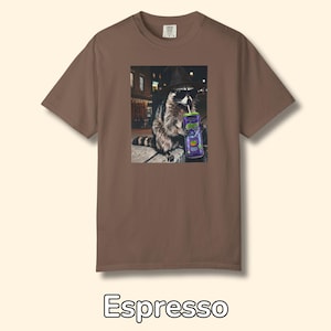 May include: A brown short-sleeve t-shirt featuring a graphic print of a raccoon wearing a black hat and sunglasses while holding a purple can with a straw. The raccoon is depicted in an outdoor setting at night. The shirt has a crew neckline.