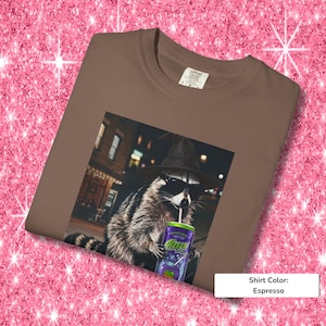 May include: An espresso-colored t-shirt featuring a graphic print of a raccoon wearing a black witch hat and sunglasses while holding a purple can with a straw. The raccoon is positioned in front of a blurred city street scene at night.