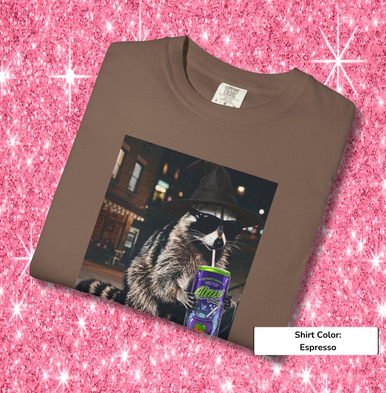 May include: An espresso-colored t-shirt featuring a graphic print of a raccoon wearing a black witch hat and sunglasses while holding a purple can with a straw. The raccoon is positioned in front of a blurred city street scene at night.