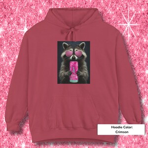 May include: A crimson hoodie featuring a graphic print of a raccoon wearing pink sunglasses and holding a pink Alani energy drink can. The hoodie has a drawstring hood and a front kangaroo pocket, showcasing a centered rectangular design.