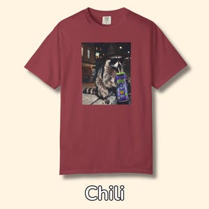 May include: A chili-colored short-sleeve t-shirt featuring a graphic print of a raccoon wearing a black fedora and sunglasses while holding a purple can with a straw. The raccoon is positioned in an urban setting at night with street lights.
