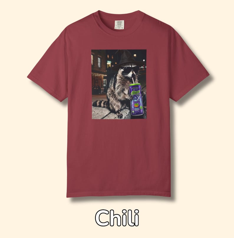 May include: A chili-colored short-sleeve t-shirt featuring a graphic print of a raccoon wearing a black fedora and sunglasses while holding a purple can with a straw. The raccoon is positioned in an urban setting at night with street lights.