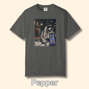 May include: A charcoal gray short-sleeve t-shirt featuring a graphic print of a raccoon wearing a black hat and sunglasses while drinking from a purple can with a straw. The raccoon is positioned in a city setting at night. The word Pepper is below.