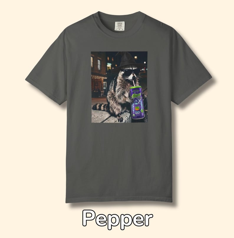May include: A charcoal gray short-sleeve t-shirt featuring a graphic print of a raccoon wearing a black hat and sunglasses while drinking from a purple can with a straw. The raccoon is positioned in a city setting at night. The word Pepper is below.