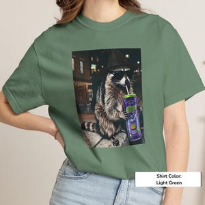 May include: A light green t-shirt featuring a graphic print of a raccoon wearing a black witch hat and sunglasses while holding a purple can with the text "Alani" and a straw. The raccoon is positioned in an outdoor urban setting at night.