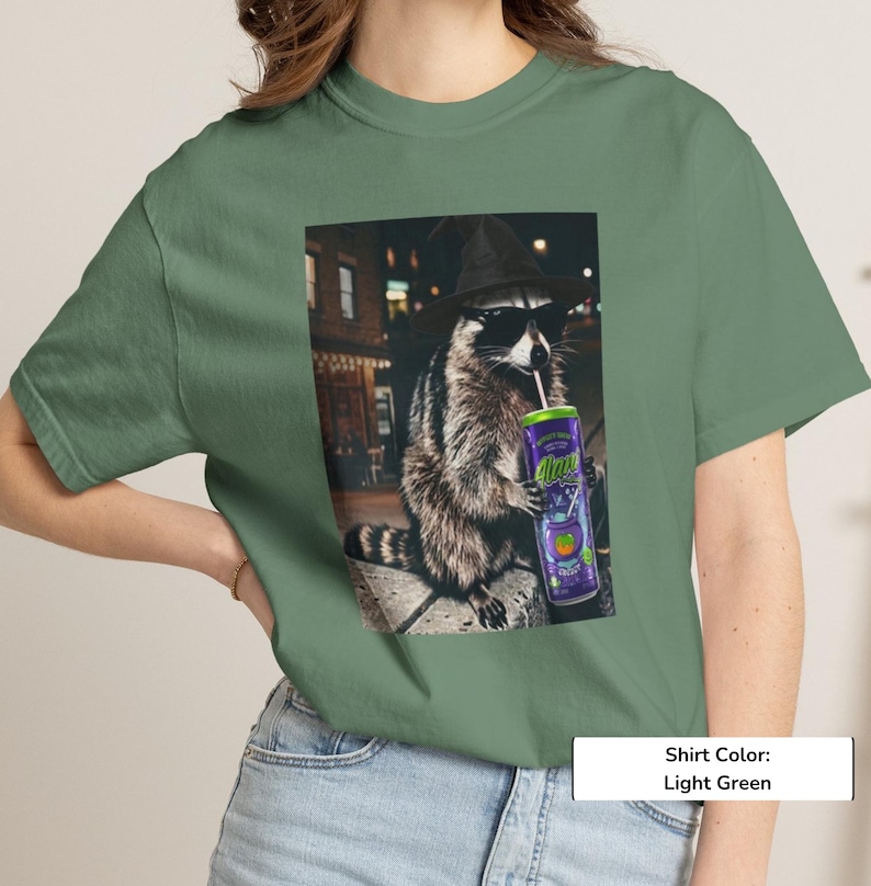 May include: A light green t-shirt featuring a graphic print of a raccoon wearing a black witch hat and sunglasses while holding a purple can with the text "Alani" and a straw. The raccoon is positioned in an outdoor urban setting at night.