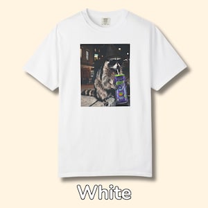 May include: A white short-sleeve t-shirt featuring a graphic print of a raccoon wearing a black hat and sunglasses while holding a purple can with a straw. The raccoon is depicted in a nighttime urban setting with blurred city lights in the background.