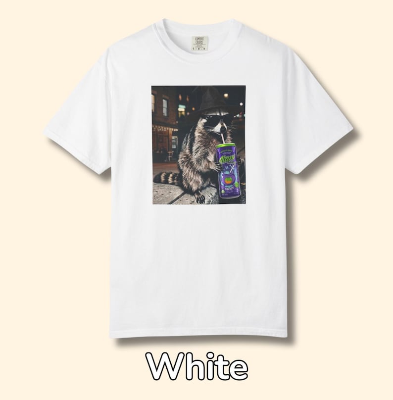 May include: A white short-sleeve t-shirt featuring a graphic print of a raccoon wearing a black hat and sunglasses while holding a purple can with a straw. The raccoon is depicted in a nighttime urban setting with blurred city lights in the background.