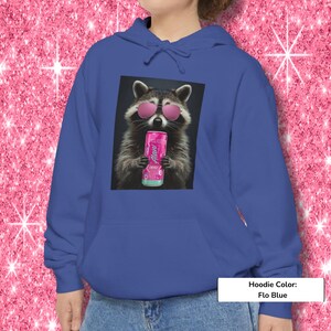 May include: A flo blue hoodie featuring a graphic print of a raccoon wearing pink sunglasses and holding a pink Alani energy drink can. The hoodie includes a drawstring hood and a front kangaroo pocket, providing a casual look for daily wear.