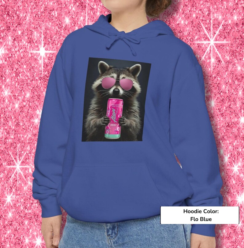 May include: A flo blue hoodie featuring a graphic print of a raccoon wearing pink sunglasses and holding a pink Alani energy drink can. The hoodie includes a drawstring hood and a front kangaroo pocket, providing a casual look for daily wear.
