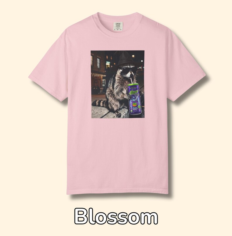 May include: A light pink short-sleeve t-shirt featuring a graphic print of a raccoon wearing a black hat and sunglasses while drinking from a purple can with a straw. The text Blossom appears below the shirt on the plain background.