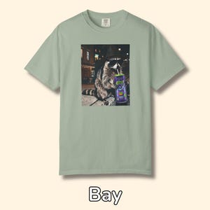 May include: A sage green short-sleeve t-shirt featuring a graphic print of a raccoon wearing a black fedora and sunglasses while holding a purple can with a straw. The raccoon is positioned in an urban setting at night with city lights visible.