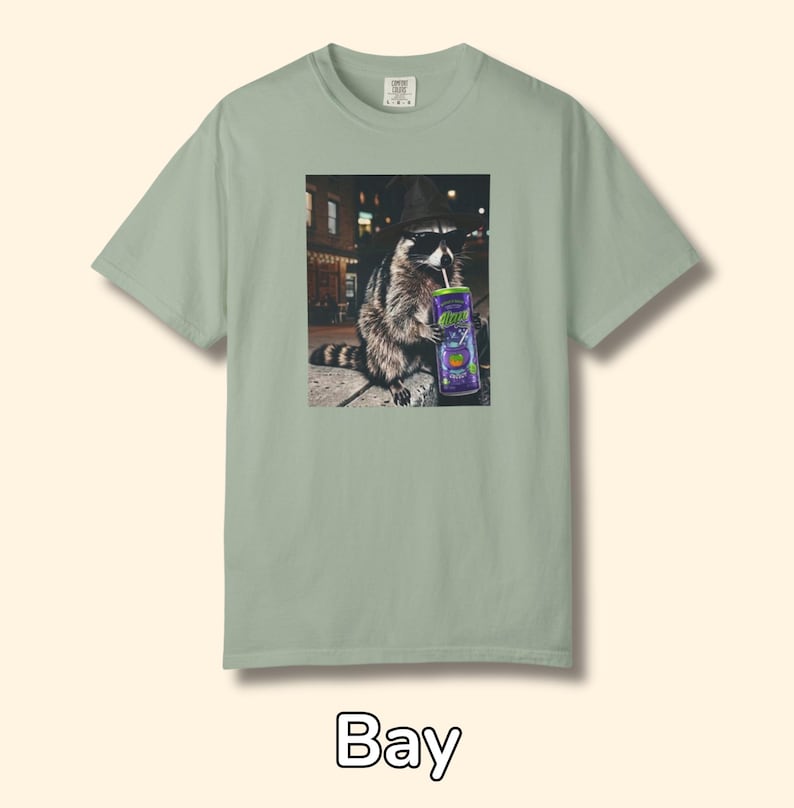May include: A sage green short-sleeve t-shirt featuring a graphic print of a raccoon wearing a black fedora and sunglasses while holding a purple can with a straw. The raccoon is positioned in an urban setting at night with city lights visible.