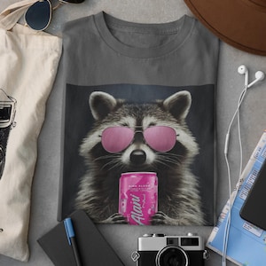 May include: A charcoal gray t-shirt featuring a graphic of a raccoon wearing pink aviator sunglasses and holding a pink can labeled Alani Nu. The raccoon has detailed fur texture and is centered on the front of the shirt, creating a bold visual.