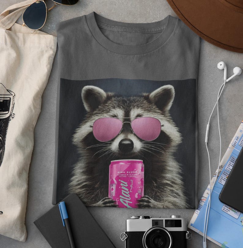 May include: A charcoal gray t-shirt featuring a graphic of a raccoon wearing pink aviator sunglasses and holding a pink can labeled Alani Nu. The raccoon has detailed fur texture and is centered on the front of the shirt, creating a bold visual.