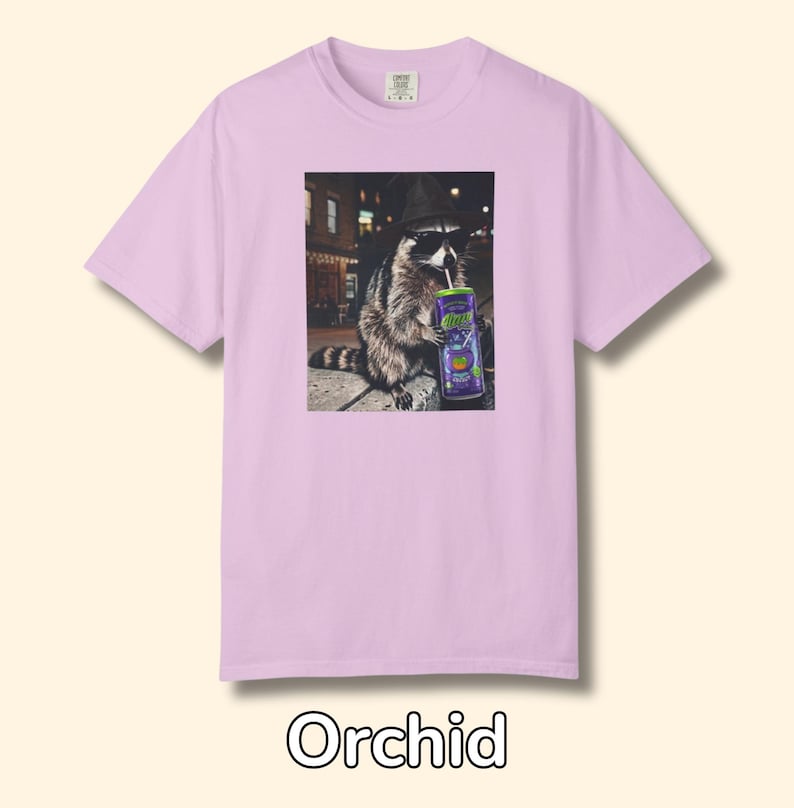 May include: An orchid-colored short-sleeve t-shirt featuring a graphic print of a raccoon wearing a black hat and sunglasses while drinking from a purple can with a straw. The raccoon is positioned in an urban setting at night with street lights.