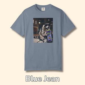 May include: A blue jean colored short-sleeve t-shirt featuring a graphic print of a raccoon wearing a black hat and sunglasses while holding a purple can with a straw. The raccoon is positioned in an urban setting at night with street lights.