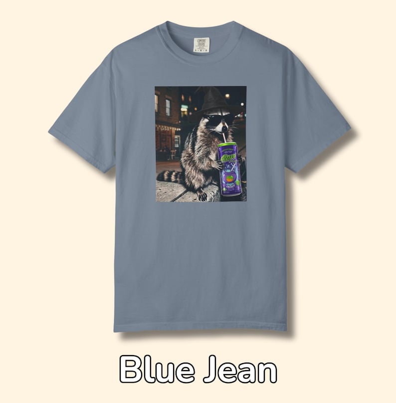 May include: A blue jean colored short-sleeve t-shirt featuring a graphic print of a raccoon wearing a black hat and sunglasses while holding a purple can with a straw. The raccoon is positioned in an urban setting at night with street lights.