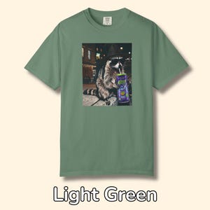 May include: A light green short-sleeve t-shirt featuring a graphic print of a raccoon wearing a black hat and sunglasses while drinking from a purple can with a straw. The raccoon is positioned in an urban setting at night with street lights.
