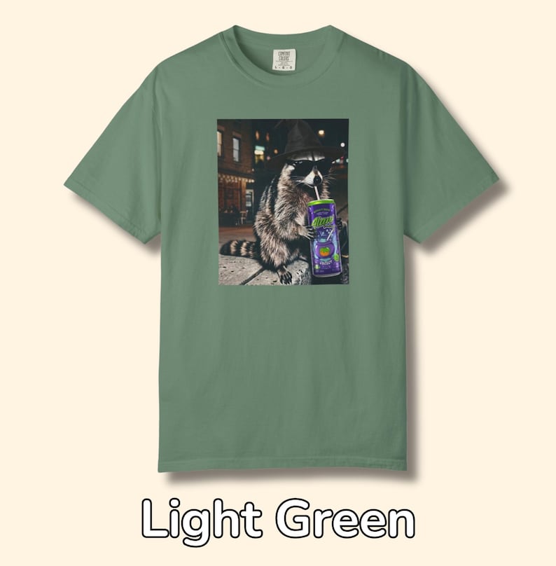 May include: A light green short-sleeve t-shirt featuring a graphic print of a raccoon wearing a black hat and sunglasses while drinking from a purple can with a straw. The raccoon is positioned in an urban setting at night with street lights.
