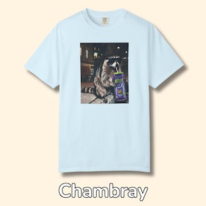 May include: A light blue short-sleeve t-shirt featuring a central graphic print of a raccoon wearing a black hat and sunglasses while holding a purple can with a straw. The raccoon is depicted in a nighttime urban setting with city lights.
