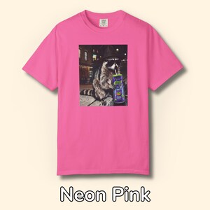 May include: A neon pink short-sleeve t-shirt featuring a graphic print of a raccoon wearing a black hat and sunglasses while drinking from a purple can labeled 'Alani' with a straw. The raccoon is positioned in an outdoor urban setting at night.
