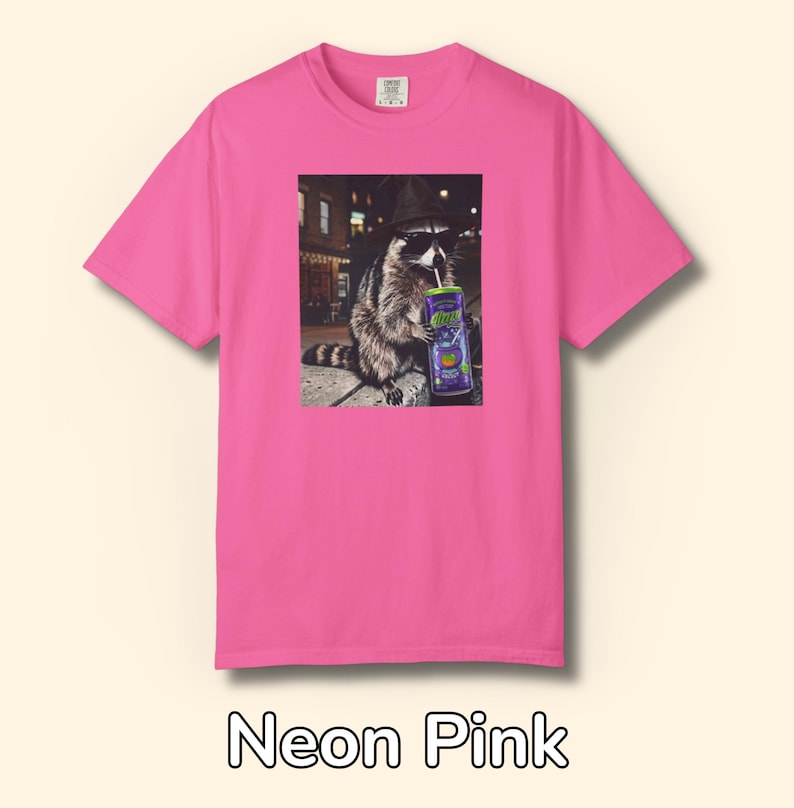 May include: A neon pink short-sleeve t-shirt featuring a graphic print of a raccoon wearing a black hat and sunglasses while drinking from a purple can labeled 'Alani' with a straw. The raccoon is positioned in an outdoor urban setting at night.