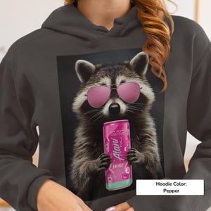 May include: A pepper-colored hoodie featuring a graphic print of a raccoon wearing pink sunglasses and holding a pink Alani Nu energy drink can. The raccoon has detailed fur, and the design is centered on the front of the hooded sweatshirt.
