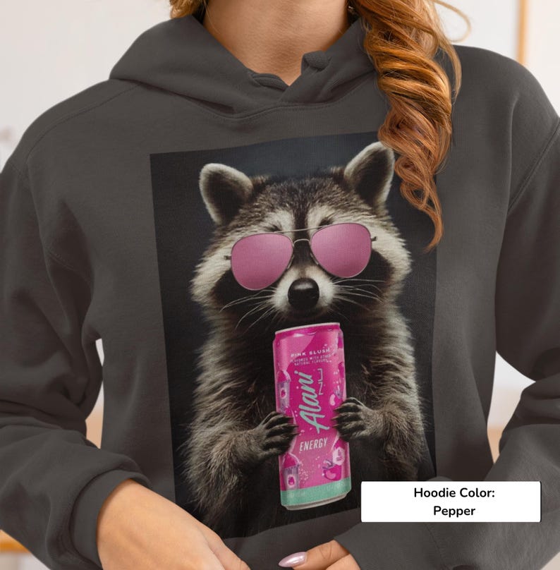 May include: A pepper-colored hoodie featuring a graphic print of a raccoon wearing pink sunglasses and holding a pink Alani Nu energy drink can. The raccoon has detailed fur, and the design is centered on the front of the hooded sweatshirt.