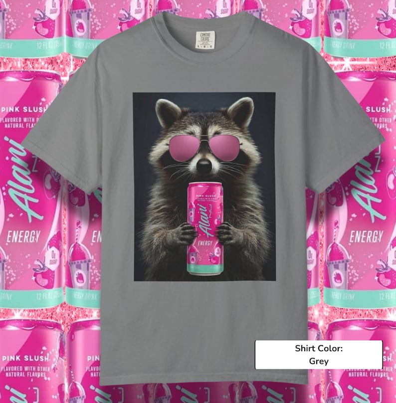 May include: A grey short-sleeve t-shirt featuring a graphic of a raccoon wearing pink sunglasses and holding a pink Alani Nu energy drink can. The design is centered on the chest, showcasing the raccoon's fur texture and the vibrant pink beverage.