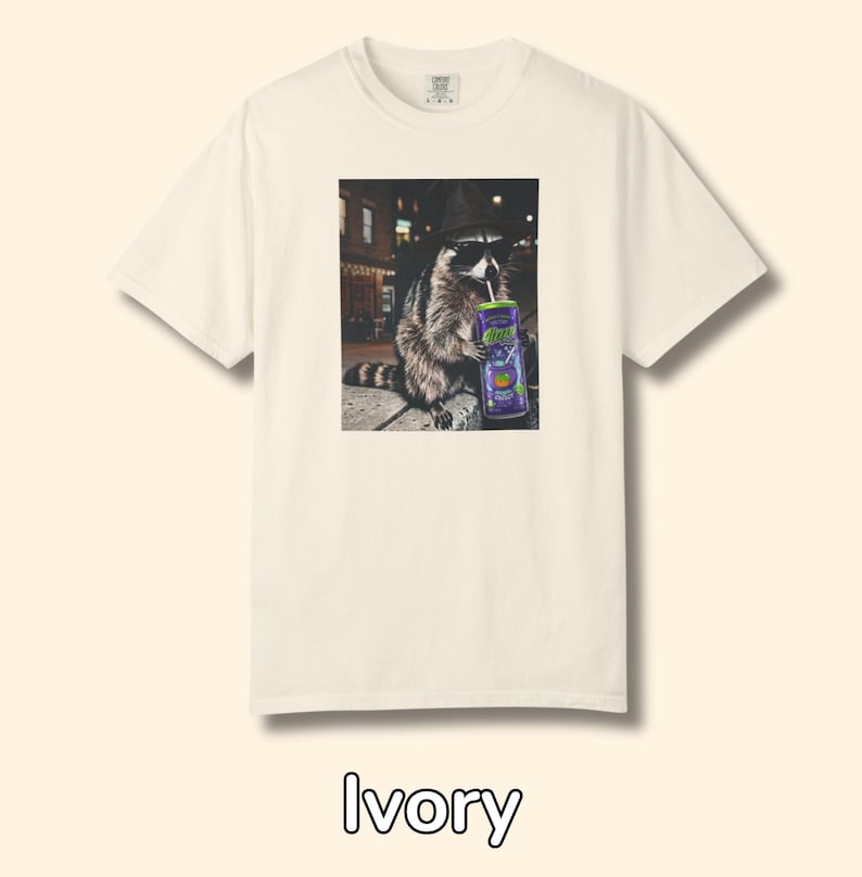 May include: An ivory short-sleeve t-shirt featuring a graphic print of a raccoon wearing a black hat and sunglasses while holding a purple can with a straw. The raccoon is positioned in a city street setting at night with blurred lights in the background.