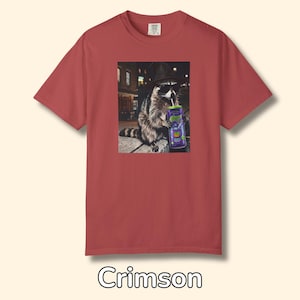 May include: A crimson short-sleeve t-shirt featuring a graphic print of a raccoon wearing a black hat and sunglasses while drinking from a purple can with a straw. The raccoon is positioned in an urban setting at night with street lights visible.