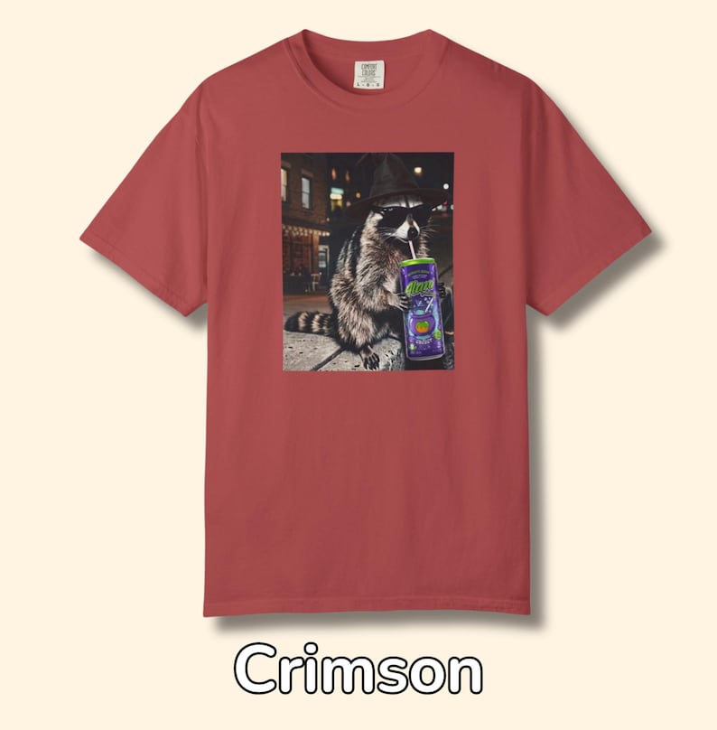 May include: A crimson short-sleeve t-shirt featuring a graphic print of a raccoon wearing a black hat and sunglasses while drinking from a purple can with a straw. The raccoon is positioned in an urban setting at night with street lights visible.