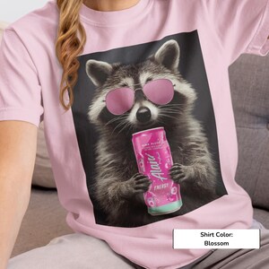 May include: A blossom pink t-shirt featuring a graphic print of a raccoon wearing pink-tinted sunglasses and holding a pink Alani Nu energy drink can. The raccoon has grey and black fur, and the design is centered on the front of the garment.