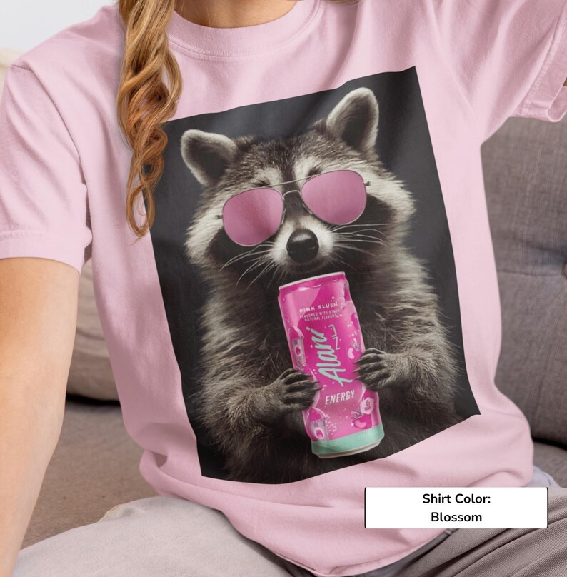 May include: A blossom pink t-shirt featuring a graphic print of a raccoon wearing pink-tinted sunglasses and holding a pink Alani Nu energy drink can. The raccoon has grey and black fur, and the design is centered on the front of the garment.