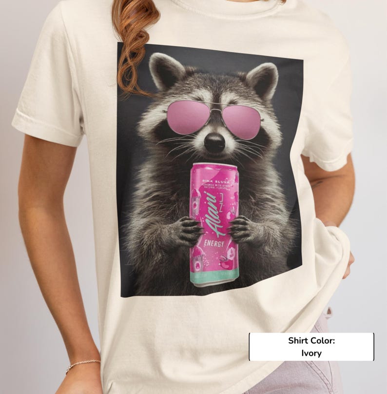 May include: An ivory t-shirt featuring a graphic print of a raccoon wearing pink aviator sunglasses and holding a pink Alani Nu energy drink can. The raccoon is centered on the shirt, which has short sleeves and a crew neckline, showcasing a detailed illustration.