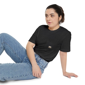 Könnte beinhalten: Dunkelgraues T-Shirt mit einer kleinen Tasche auf der Brust. Die Tasche hat ein detailliertes Stickdesign. Das Model trägt eine blaue Jeans. Das Shirt ist ein kurzärmeliges Rundhals-Shirt.