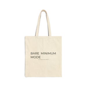 May include: A beige tote bag with black text that reads "BARE MINIMUM MODE" and "running on low effort". The bag has a simple design with two handles and is made of a canvas-like material.