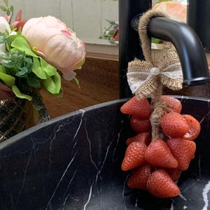 May include: A decorative soap shaped like strawberries hangs from a black faucet. The soap is tied with twine and a burlap bow. A bouquet of pink and white flowers sits in a gold vase near a black marble sink.