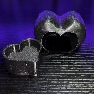 May include: Two black heart-shaped objects, one solid and the other a hollow mould, are on a dark surface. The objects appear 3D-printed, with visible layer lines. The larger heart has a cutout, and the smaller one is a solid container.