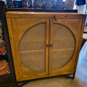 Puede incluir: Armario de madera clara con dos puertas, cada una con un panel redondo de ratán tejido. El armario tiene patas de metal negro. Se ven tazas y teteras decorativas en la parte superior.